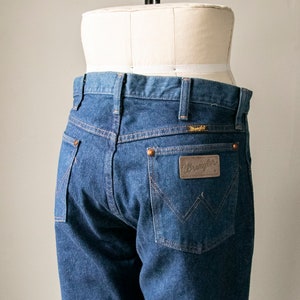 Puede incluir: Un par de pantalones vaqueros azules con un parche de cuero marr&oacute;n de Wrangler en el bolsillo trasero.
