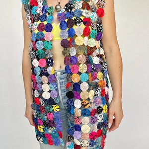 Peut inclure: Un gilet sans manches composé de nombreuses petites pièces de tissu circulaires en forme de yo-yos, déclinées dans une grande variété de couleurs et de motifs. Le vêtement présente une ouverture sur le devant, avec des éléments assemblés pour créer un aspect patchwork texturé, semi-transparent et multicolore.