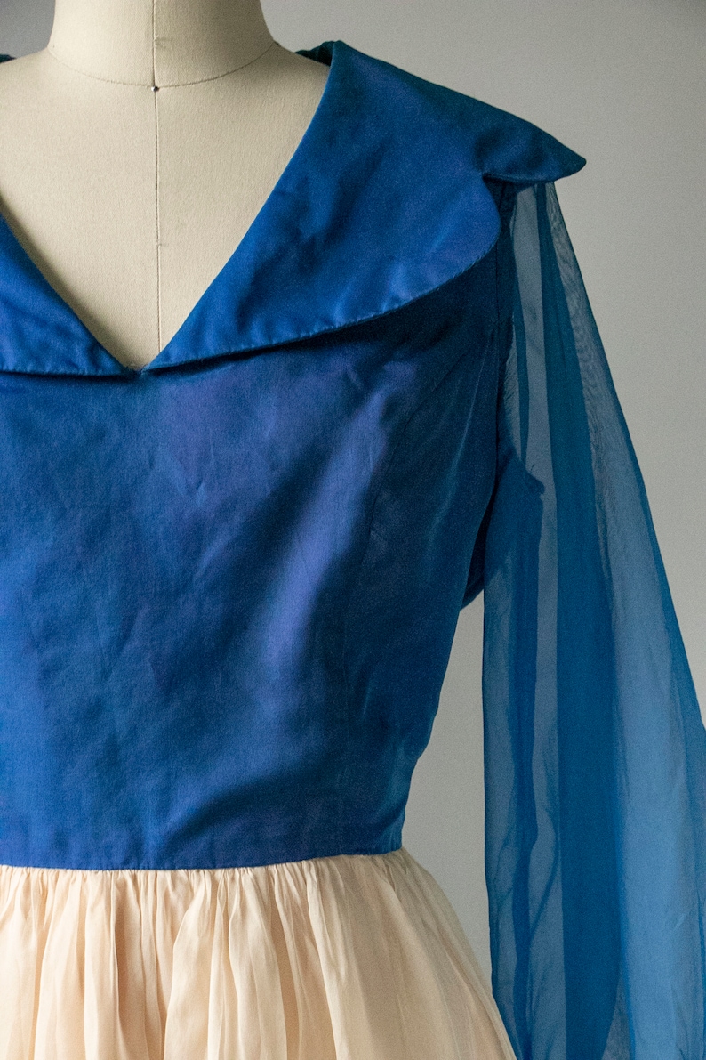 May include: A blue dress with a sheer overlay and a large collar. The dress has a fitted bodice and a full skirt.