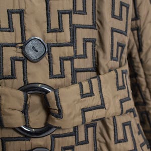 May include: A brown quilted jacket with a black geometric pattern and a black belt. The jacket has two black buttons and a black ring buckle.