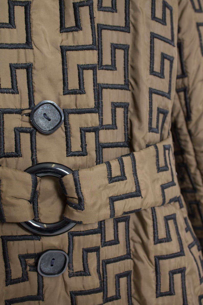 May include: A brown quilted jacket with a black geometric pattern and a black belt. The jacket has two black buttons and a black ring buckle.