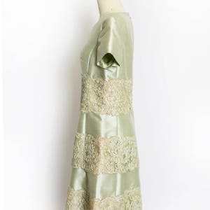 May include: A pale green dress with a fitted bodice and a flared skirt. The dress is adorned with delicate lace trim around the neckline, sleeves, and hem.