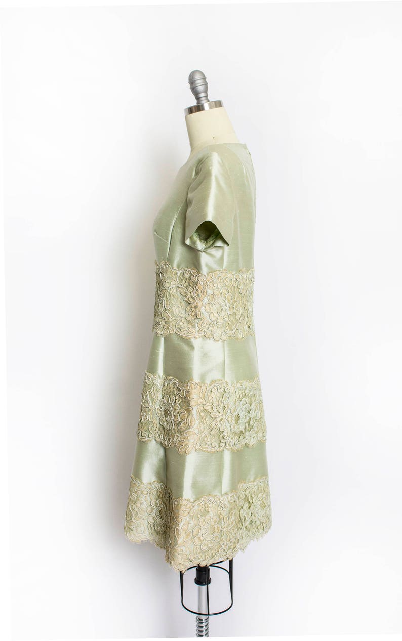 May include: A pale green dress with a fitted bodice and a flared skirt. The dress is adorned with delicate lace trim around the neckline, sleeves, and hem.