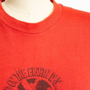 May include: Red crew neck t-shirt with a graphic design. The design features the text "AND ON THE EIGHTH DAY" around a globe with a lightning bolt and a radio.