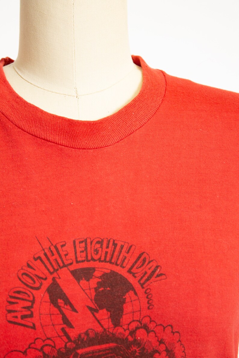May include: Red crew neck t-shirt with a graphic design. The design features the text "AND ON THE EIGHTH DAY" around a globe with a lightning bolt and a radio.