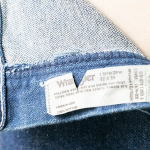 Puede incluir: Un primer plano de una etiqueta de mezclilla azul Wrangler con texto blanco. La etiqueta dice "13MWZPW 32 X 34" e incluye instrucciones de lavado. La etiqueta est&aacute; unida al interior del jean.
