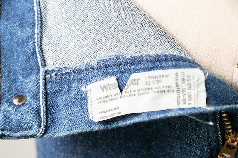 Puede incluir: Un primer plano de una etiqueta de mezclilla azul Wrangler con texto blanco. La etiqueta dice "13MWZPW 32 X 34" e incluye instrucciones de lavado. La etiqueta est&aacute; unida al interior del jean.