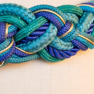 Pu&ograve; includere: Cintura intrecciata nelle tonalit&agrave; del verde acqua, turchese e blu navy, rifinita con bordi dorati. La cintura &egrave; realizzata in corda spessa e intrecciata ed &egrave; esposta su un manichino. Un accessorio elegante.