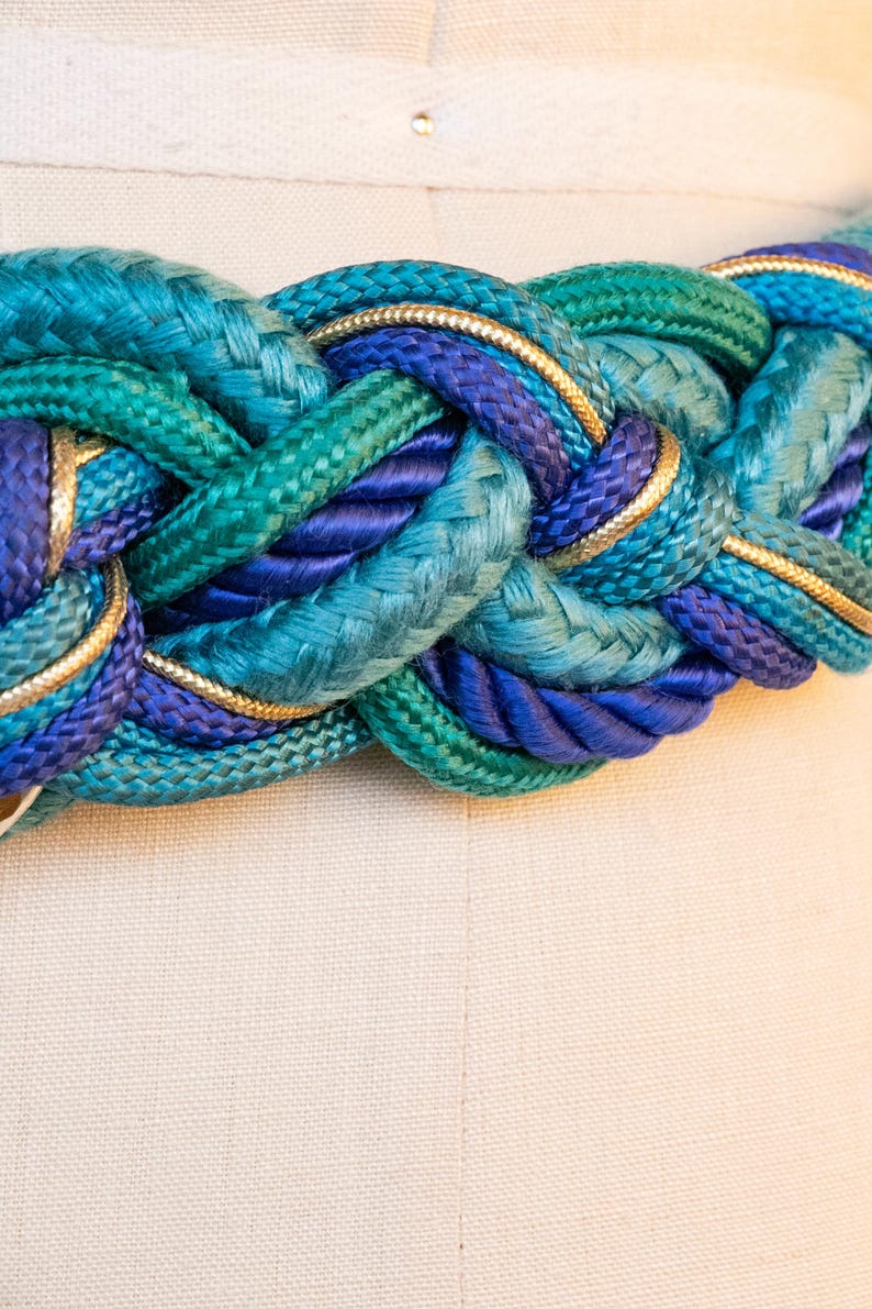 Pu&ograve; includere: Cintura intrecciata nelle tonalit&agrave; del verde acqua, turchese e blu navy, rifinita con bordi dorati. La cintura &egrave; realizzata in corda spessa e intrecciata ed &egrave; esposta su un manichino. Un accessorio elegante.