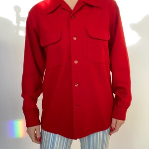 Pode incluir: Uma camisa vermelha brilhante de manga comprida com dois bolsos no peito e uma frente abotoada. A camisa tem uma gola clássica e é feita de um material liso. As mangas são compridas e abotoadas nos punhos. A camisa é combinada com calças listradas azuis e brancas.