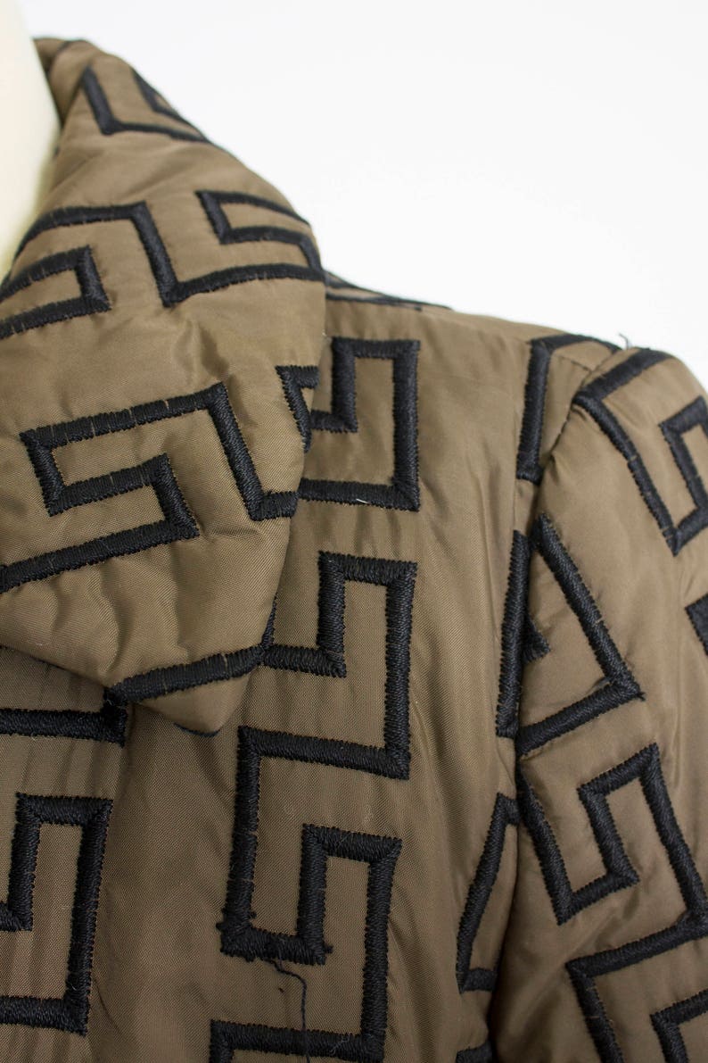 May include: A brown quilted jacket with a black geometric pattern. The jacket has a high collar and a zip closure.