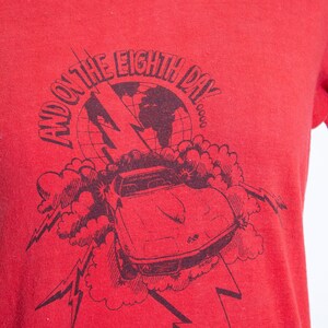 May include: Red t-shirt with a black graphic featuring a car, lightning bolts, and the text "AND ON THE EIGHTH DAY". The design includes a globe and cloud-like shapes.