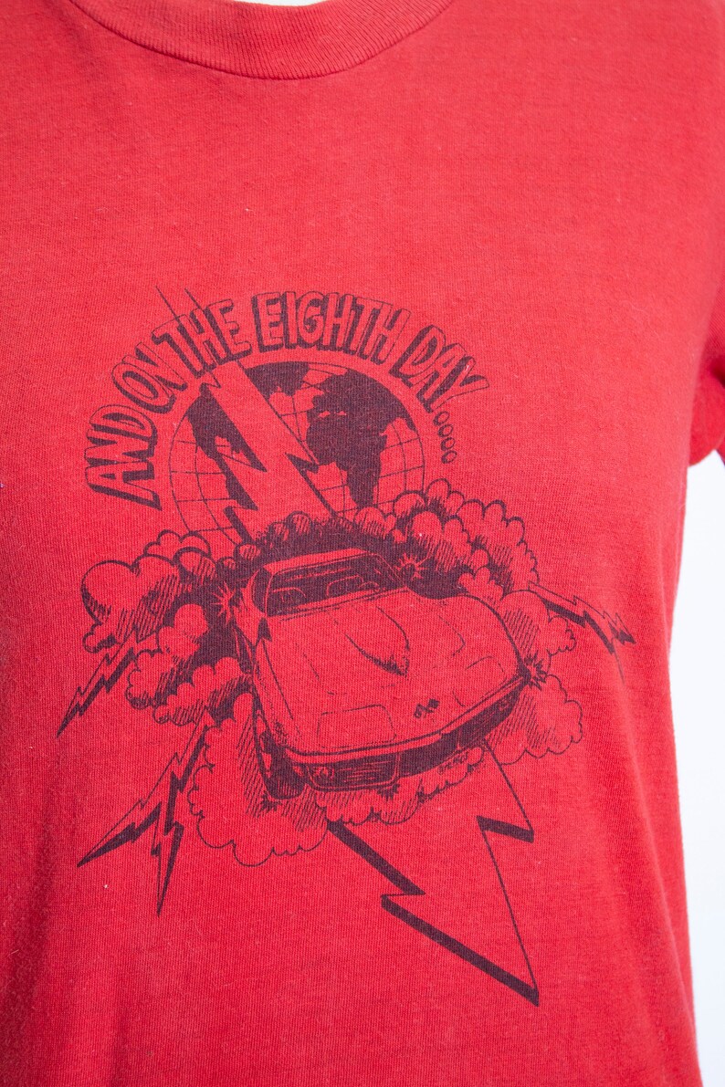 May include: Red t-shirt with a black graphic featuring a car, lightning bolts, and the text "AND ON THE EIGHTH DAY". The design includes a globe and cloud-like shapes.
