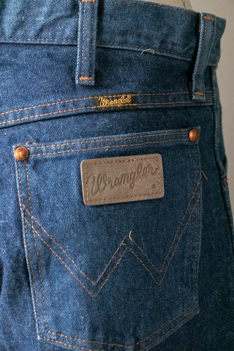 Puede incluir: Un par de pantalones vaqueros azules con un parche de cuero marr&oacute;n en el bolsillo trasero que dice "Wrangler".