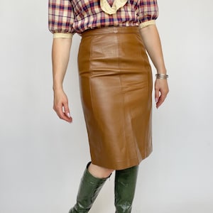 May include: A brown leather pencil skirt paired with a plaid blouse and green knee-high boots. The skirt is knee-length and has vertical seams. The blouse has a ruffled collar and sleeves. The boots are a glossy green.