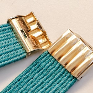 Pu&ograve; includere: Primo piano di una fascia elastica turchese con chiusure in metallo color oro. Le chiusure hanno un design a coste e il testo "SHOE" &egrave; visibile su una. La fascia &egrave; realizzata in materiale tessuto.