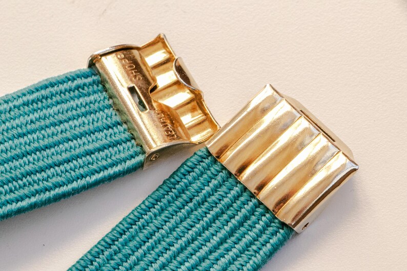 Pu&ograve; includere: Primo piano di una fascia elastica turchese con chiusure in metallo color oro. Le chiusure hanno un design a coste e il testo "SHOE" &egrave; visibile su una. La fascia &egrave; realizzata in materiale tessuto.