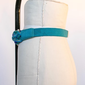 Pu&ograve; includere: Una cintura turchese con un nodo decorativo, esposta su un manichino. Il nodo presenta accenti blu, gialli e dorati. La cintura &egrave; realizzata in materiale tessuto.