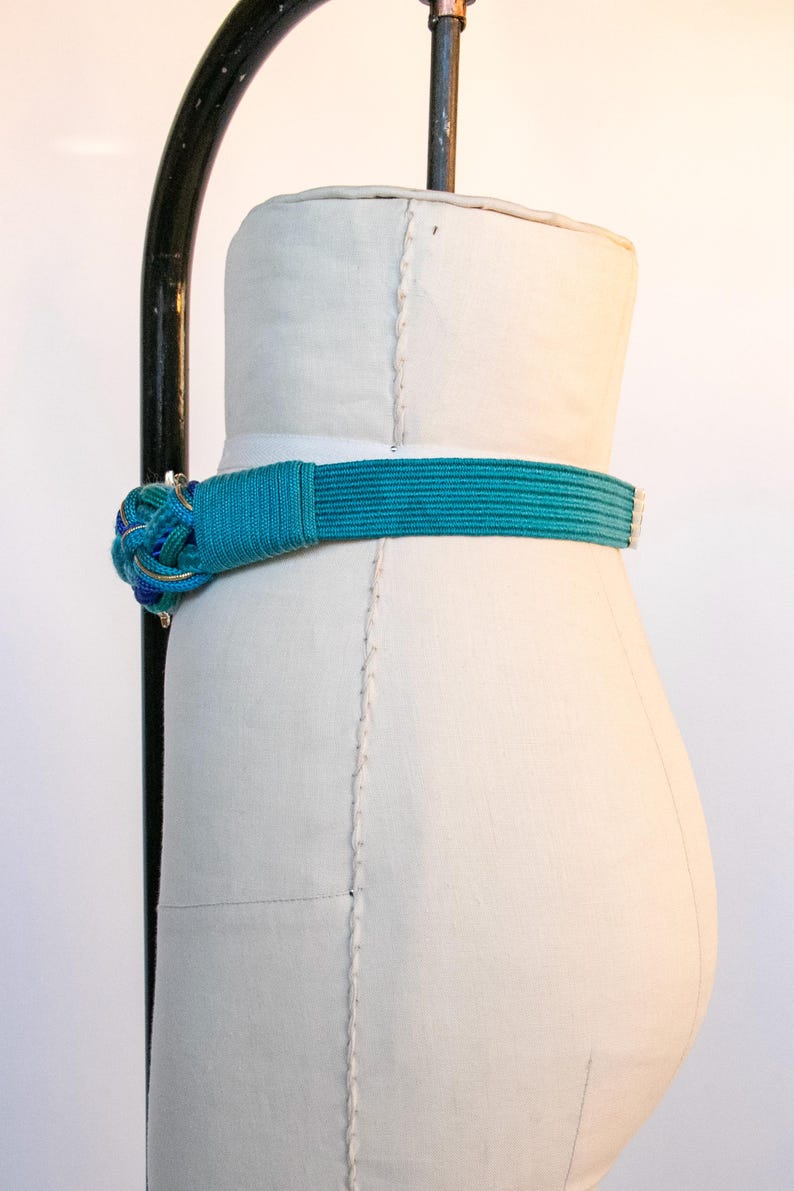 Pu&ograve; includere: Una cintura turchese con un nodo decorativo, esposta su un manichino. Il nodo presenta accenti blu, gialli e dorati. La cintura &egrave; realizzata in materiale tessuto.