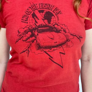 May include: Red t-shirt with a black graphic design. The design features a car, lightning bolts, clouds, and the text "AND ON THE EIGHTH DAY". The shirt is made of a soft material.