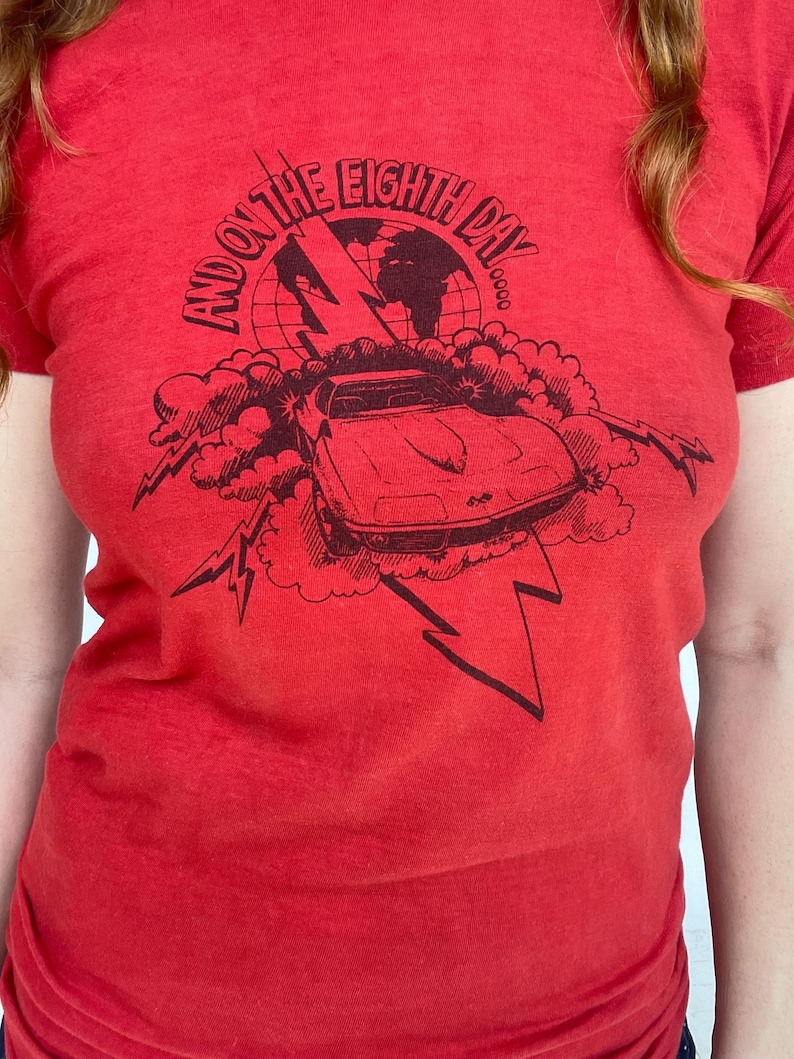 May include: Red t-shirt with a black graphic design. The design features a car, lightning bolts, clouds, and the text "AND ON THE EIGHTH DAY". The shirt is made of a soft material.