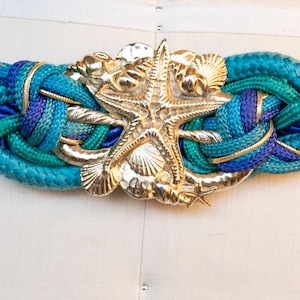 Pu&ograve; includere: Una cintura intrecciata con fibbia a forma di stella marina color oro e dettagli a conchiglia. La cintura presenta un mix di corda blu e turchese, con dettagli color oro. Un accessorio di moda a tema nautico.