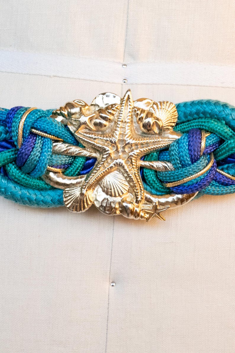 Pu&ograve; includere: Una cintura intrecciata con fibbia a forma di stella marina color oro e dettagli a conchiglia. La cintura presenta un mix di corda blu e turchese, con dettagli color oro. Un accessorio di moda a tema nautico.