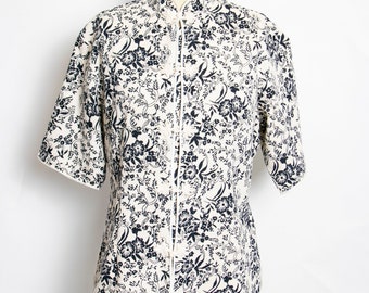 May include: Cream-colored short-sleeved shirt with a mandarin collar and a floral pattern in navy blue. The shirt is displayed on a mannequin. The shirt has a white trim and a button-up front.
