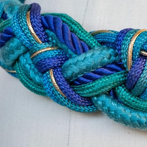 Pu&ograve; includere: Cintura in corda intrecciata nei toni del verde acqua, turchese e blu navy, rifinita con bordi color oro. La cintura &egrave; realizzata in corda spessa e intrecciata ed &egrave; esposta su un manichino.