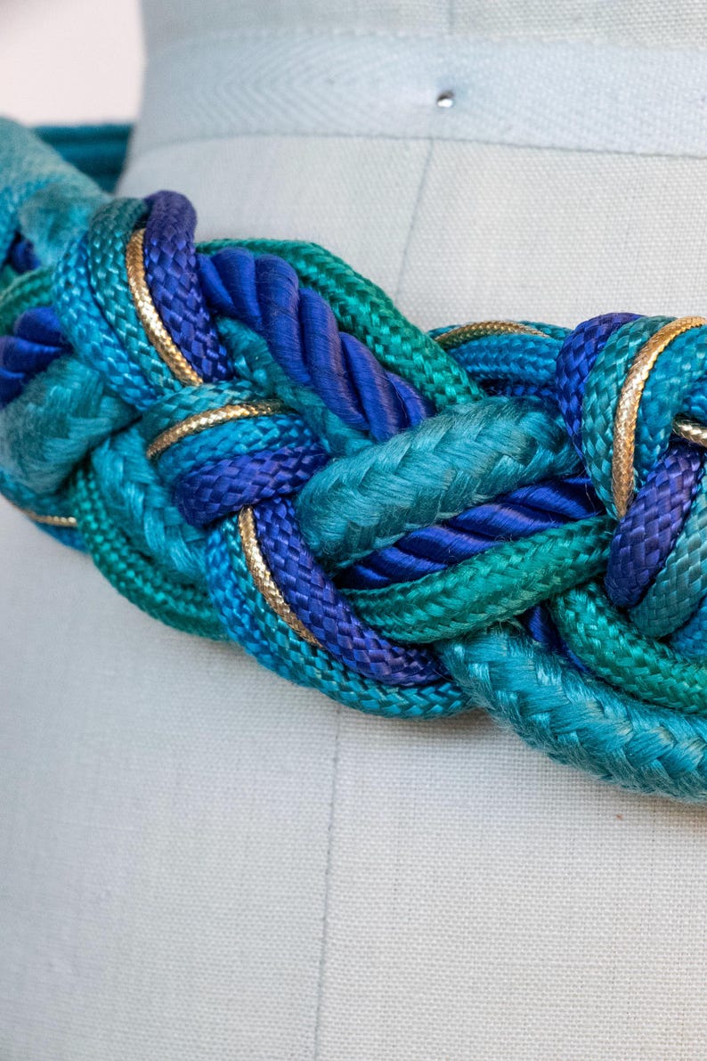 Pu&ograve; includere: Cintura in corda intrecciata nei toni del verde acqua, turchese e blu navy, rifinita con bordi color oro. La cintura &egrave; realizzata in corda spessa e intrecciata ed &egrave; esposta su un manichino.