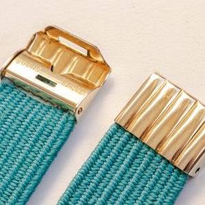 Pu&ograve; includere: Primo piano di una cintura elastica turchese con chiusure in metallo color oro. Le chiusure hanno una forma rettangolare con un design a coste. La cintura &egrave; realizzata in materiale tessuto.