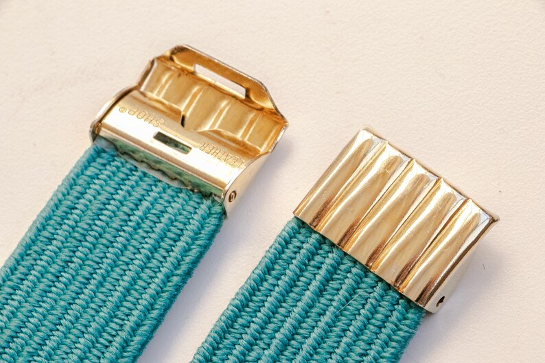 Pu&ograve; includere: Primo piano di una cintura elastica turchese con chiusure in metallo color oro. Le chiusure hanno una forma rettangolare con un design a coste. La cintura &egrave; realizzata in materiale tessuto.