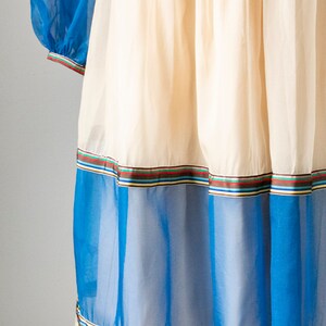 May include: A white dress with blue and white sheer panels and a multi-colored ribbon trim. The dress has a gathered skirt and a fitted bodice.