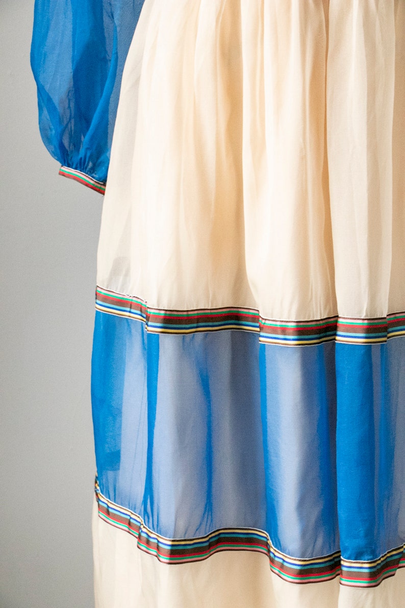 May include: A white dress with blue and white sheer panels and a multi-colored ribbon trim. The dress has a gathered skirt and a fitted bodice.
