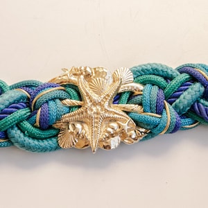 Pu&ograve; includere: Cintura intrecciata nei toni del verde acqua, blu e oro. La cintura presenta una fibbia a forma di stella marina color oro. Questa cintura a tema nautico &egrave; un accessorio elegante.