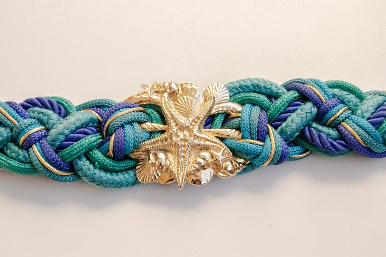 Pu&ograve; includere: Cintura intrecciata nei toni del verde acqua, blu e oro. La cintura presenta una fibbia a forma di stella marina color oro. Questa cintura a tema nautico &egrave; un accessorio elegante.