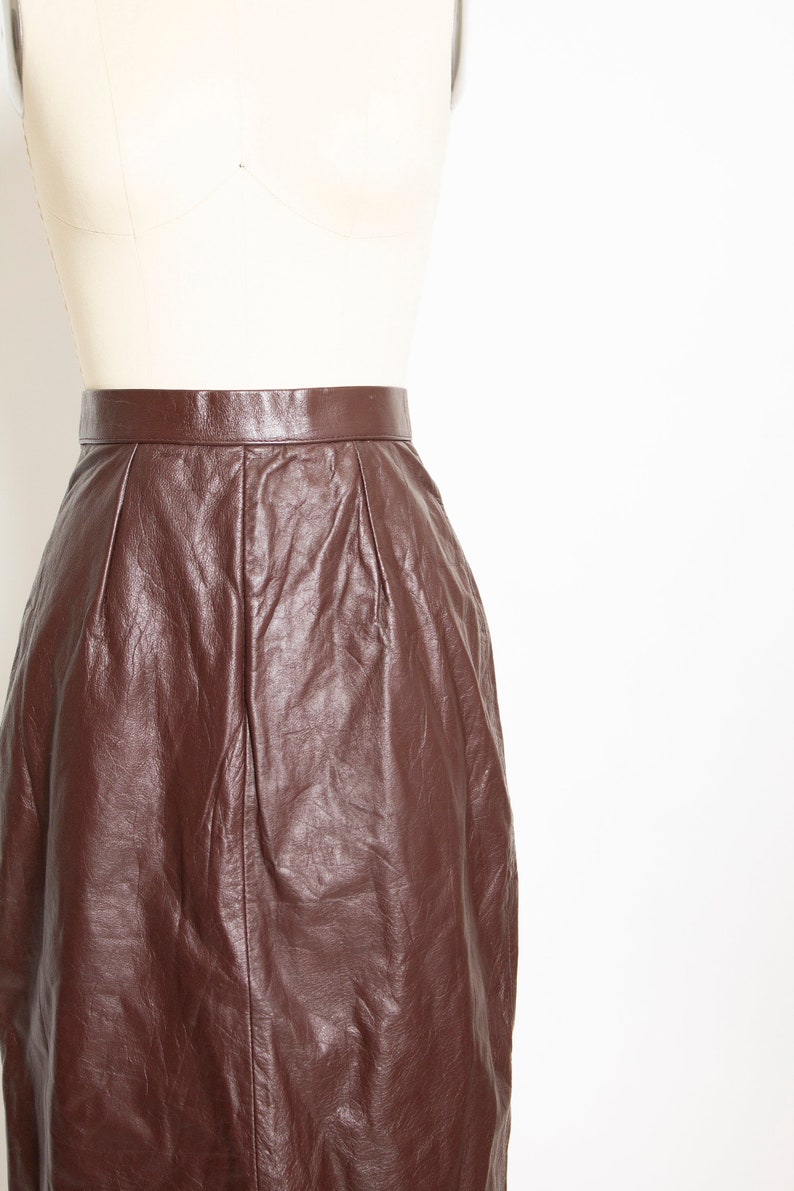 May include: A dark brown leather skirt displayed on a mannequin. The skirt has a high waist and a knee-length hemline. The leather has a slightly wrinkled texture, and the skirt appears to be well-made.