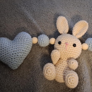 Può includere: Clip per ciuccio per bambini fatto a mano all'uncinetto. Presenta un ciuccio azzurro, un coniglietto color crema, due cuori azzurri e perline di legno. Il coniglietto ha occhi neri e naso rosa. Su tessuto grigio.