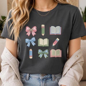 Könnte beinhalten: Ein dunkelgraues T-Shirt mit einem Grafikdruck aus drei Schleifen in rosa und blau karierten Mustern, drei offenen Büchern mit hellen Seiten und drei Stiften in Grün, Blau und Rosa, die in einem gitterartigen Muster angeordnet sind.