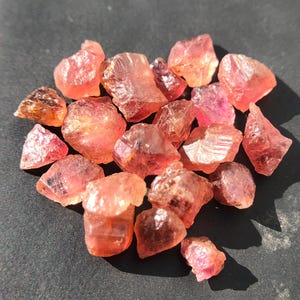 Könnte beinhalten: Eine Ansammlung roher, ungeschliffener Edelsteine in Rosa- und Orangetönen. Die Steine, von unterschiedlicher Größe und Form, weisen eine durchscheinende Qualität auf. Sie liegen auf einer dunklen Oberfläche, wobei sich das Licht in ihren Facetten spiegelt.