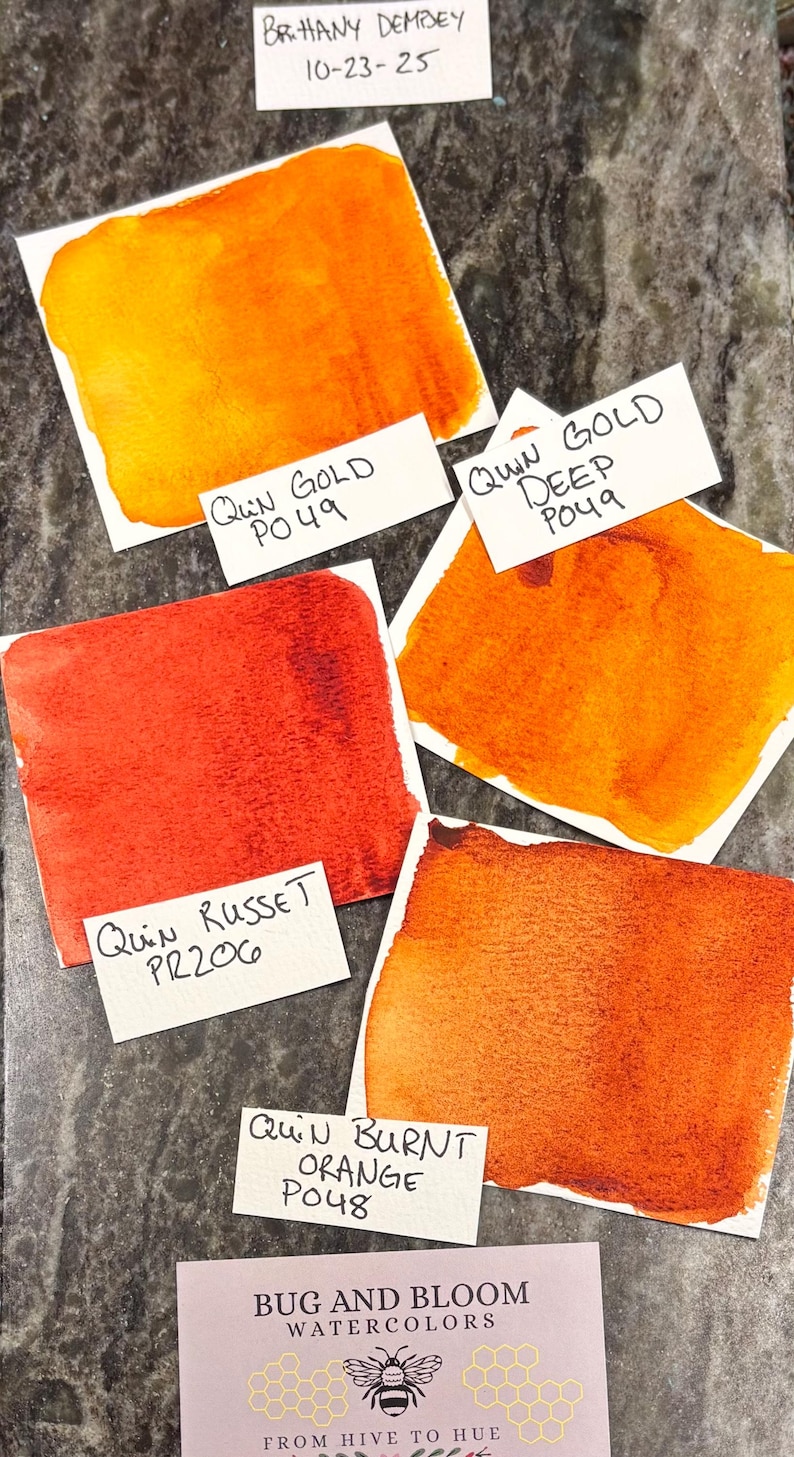 May include: Watercolor paint swatches in shades of orange and red, including Quin Gold, Quin Gold Deep, Quin Russet, and Quin Burnt Orange. Each swatch is labeled with the paint name and code. The image also includes the text "Bug and Bloom Watercolors."