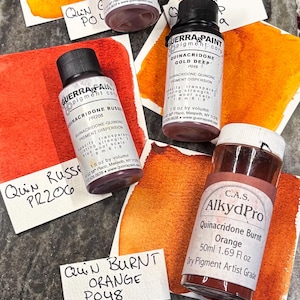 May include: An assortment of watercolor pigment bottles and swatches in shades of orange and red. The bottles are labeled with pigment names like Quinacridone Gold and Quinacridone Burnt Orange. The image also includes a card that says "Bug and Bloom Watercolors".