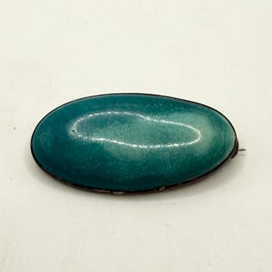 May include: An oval-shaped brooch featuring a smooth, polished teal-colored stone with subtle white marbling and a crackled texture. The stone is set within a dark, thin metal frame that follows the curved perimeter of the oval shape.