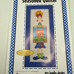 May include: A sewing pattern booklet titled Seasoned Quilter by Amy Bradley Designs. The cover features a cartoon character holding a sign that says Welcome. It includes instructions for a 23 inch by 51 inch wall-hanging quilt project.