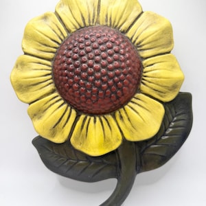 May include: A decorative wall plaque shaped like a sunflower with bright yellow petals, a textured dark reddish-brown center, and a dark green stem with two leaves. The piece features a molded, three-dimensional design with visible shading.