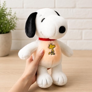 Può includere: Un peluche bianco del personaggio Snoopy con orecchie nere, naso nero e un collare rosso. Il giocattolo presenta un pannello pettorale color arancione chiaro con un ricamo giallo del personaggio Woodstock al centro del petto.