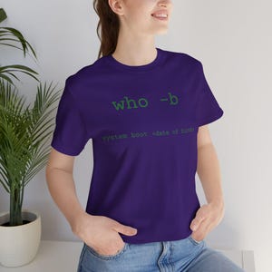 May include: A purple t-shirt with green text that reads "who -b" above "system boot <date of birth>". The shirt is a crew neck style and is made of a soft material.