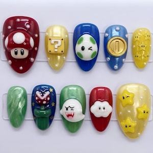 May include: A set of ten artificial nails with various designs inspired by the Super Mario video game series. The nails feature characters and symbols like a mushroom, question mark block, coin, and stars, in a variety of colors including red, yellow, blue, and green.