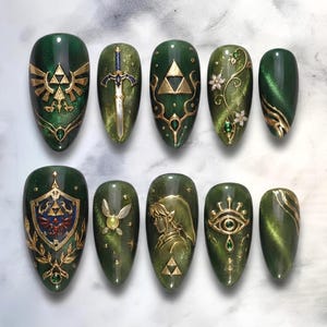 May include: A set of ten almond-shaped press-on nails featuring a deep forest green cat-eye magnetic polish base. The designs include gold-toned emblems, a sword, a shield, a fairy, a profile silhouette, and floral accents with small gems.