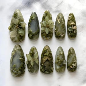 May include: A set of ten almond-shaped press-on nails featuring a forest-inspired theme with shades of sage green, moss, and grey. The designs include 3D elements like a flower, a butterfly, a small arched door, tiny mushrooms, and clear dew drops.